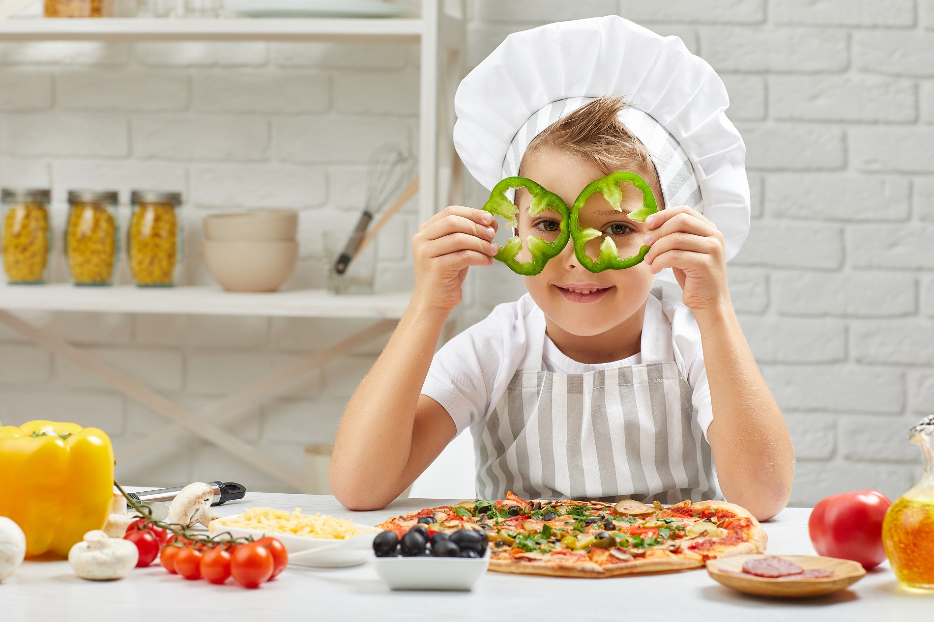 little boy in chef hat and an apron cooking pizza in the kitchen. the child holding green bell peppers . having fun