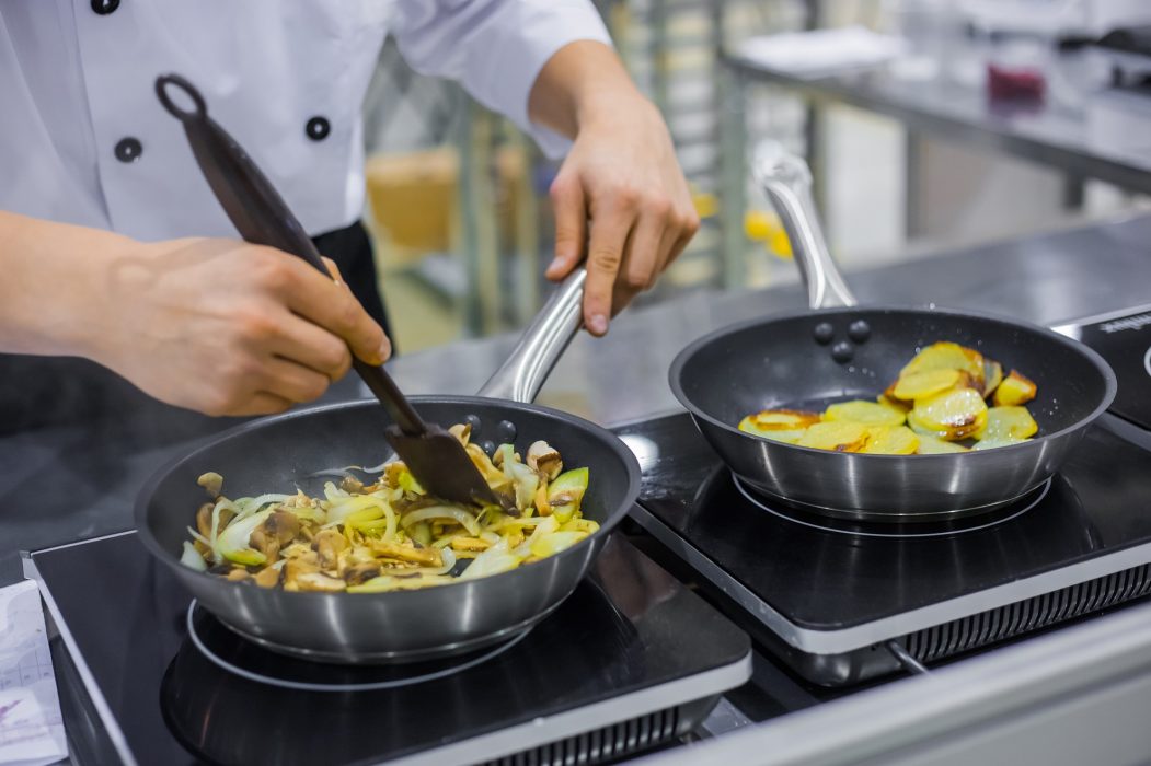 Chef cooking sliced champignon mushrooms, onions and potatoes in frying pans with oil on electric stove at cuisine of restaurant. Professional cooking, catering, cookery, gastronomy and food concept